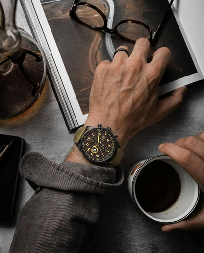 Person wearing a detailed wristwatch, holding a cup of coffee, with a book and glasses in the background.