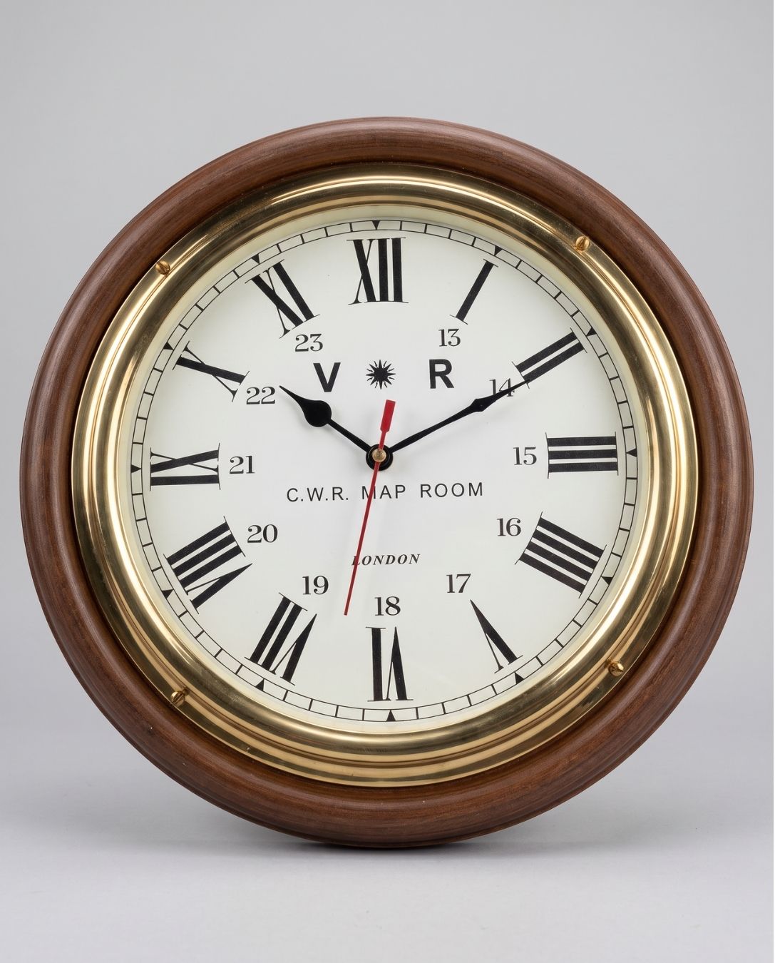 A round wall clock with a dark brown wooden frame, polished brass bezel, and an off-white clock face featuring black Roman numerals and hands. The brand 'C.