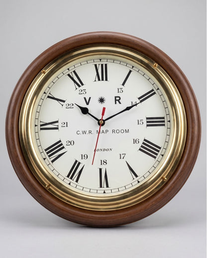 A round wall clock with a dark brown wooden frame, polished brass bezel, and an off-white clock face featuring black Roman numerals and hands. The brand 'C.