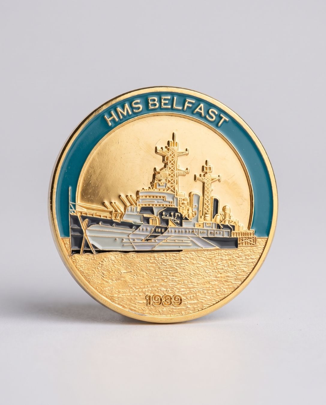 A round commemorative coin or medallion for HMS Belfast, featuring a gold-toned ship design with a teal-blue outer ring and the text 'HMS BELFAST' at the top, photographed on a light gray background.