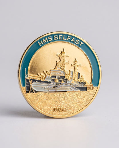 A round commemorative coin or medallion for HMS Belfast, featuring a gold-toned ship design with a teal-blue outer ring and the text 'HMS BELFAST' at the top, photographed on a light gray background.
