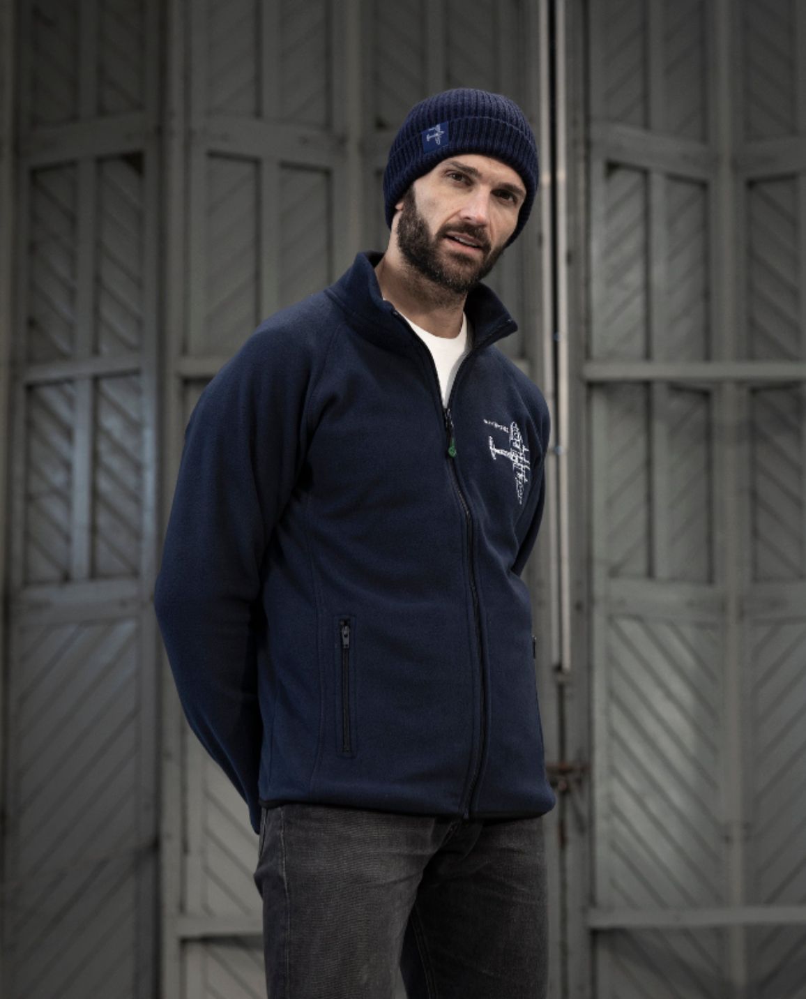 Man looking at camera wearing Navy Blueprint Spitfire Fleece with a White Spitfire Blueprint design on the left chest area, and also wearing a Navy Blue Spitfire Cap with the Spitfire Blueprint design embossed on it.