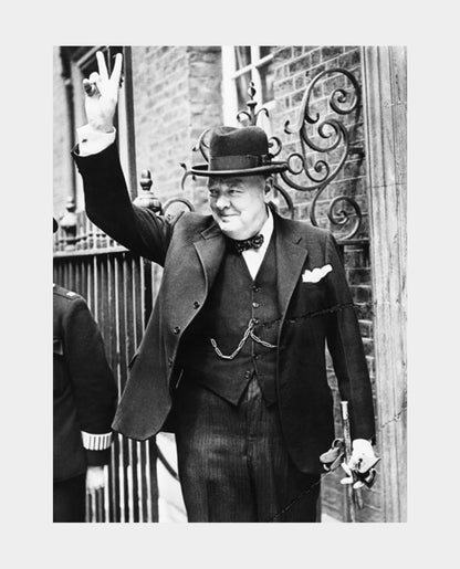 A black and white photograph of Winston Churchill in a suit and hat, raising his right hand in a 'V' shape, with a decorative wall in the background.