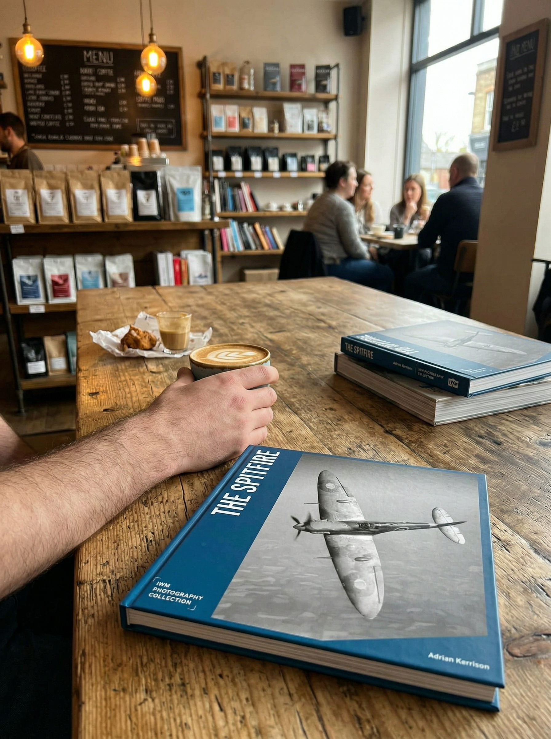 Person holding a coffee cup with a book titled 'The Spitfire' on a wooden table in a cafe.