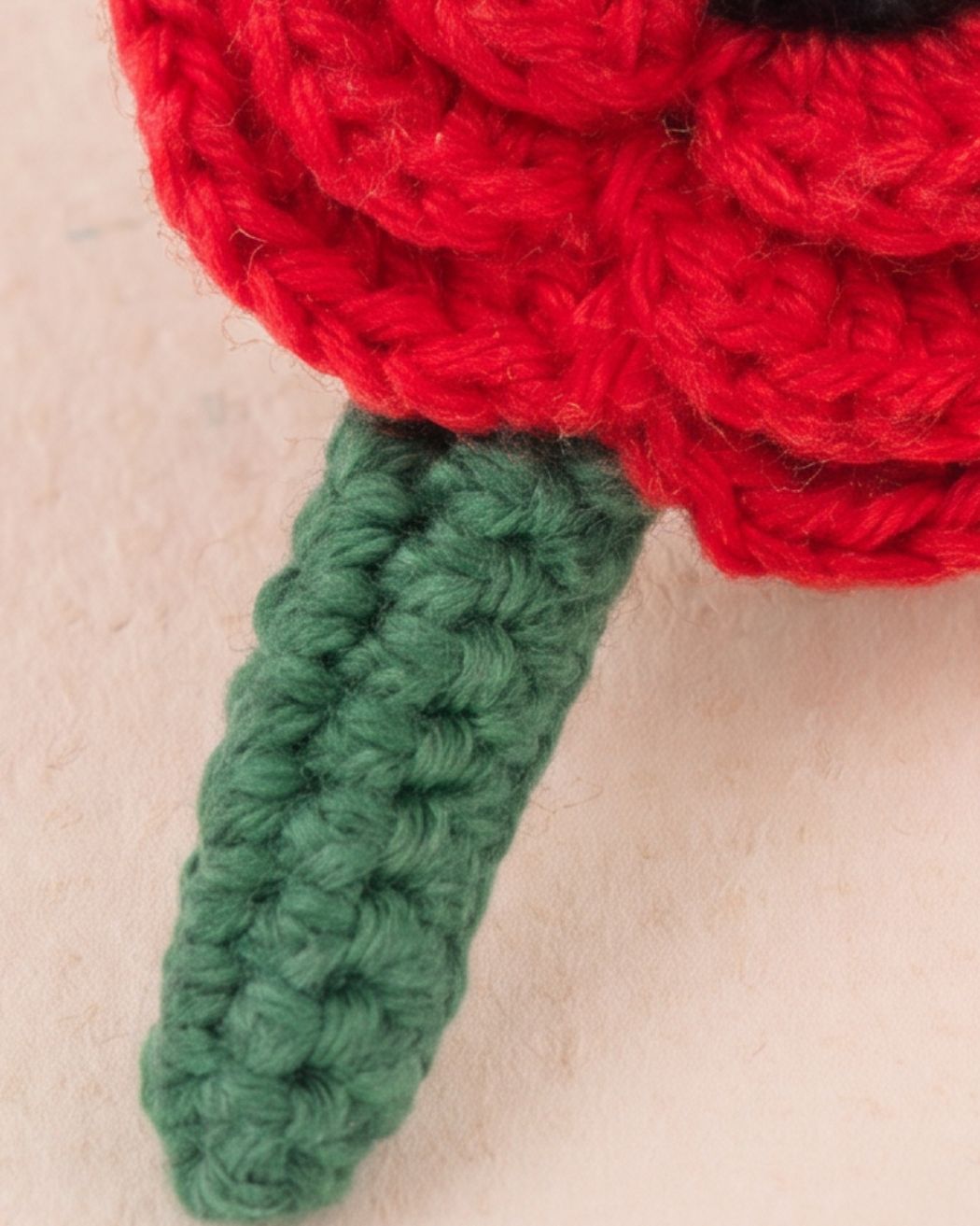 Close-up of a crocheted flower with a bright red textured bloom and a green stem on a light beige background.